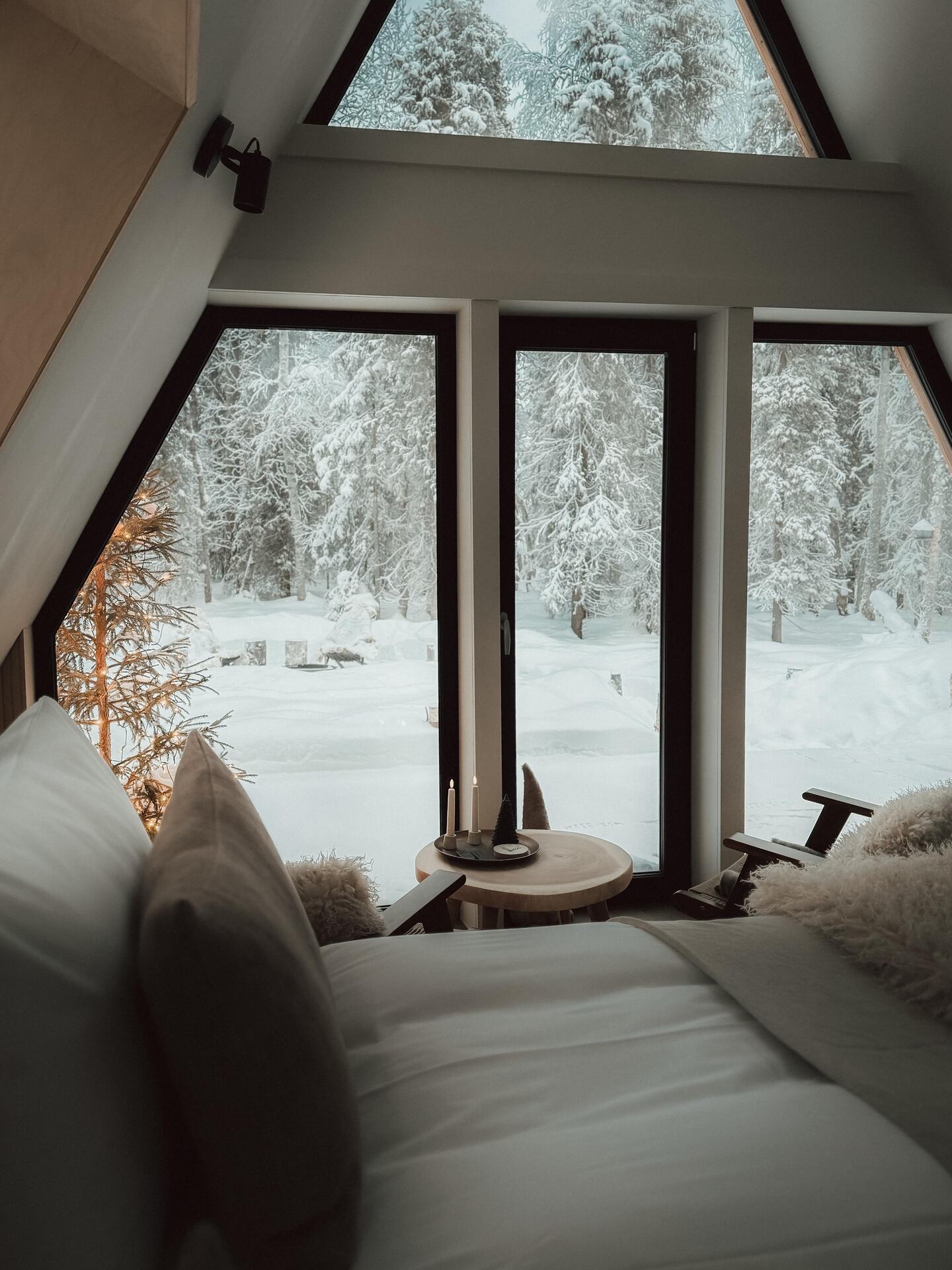 The view from a cabin bed at Sixty Eight North through tall A-frame windows onto snow-laden pines in pale winter daylight.