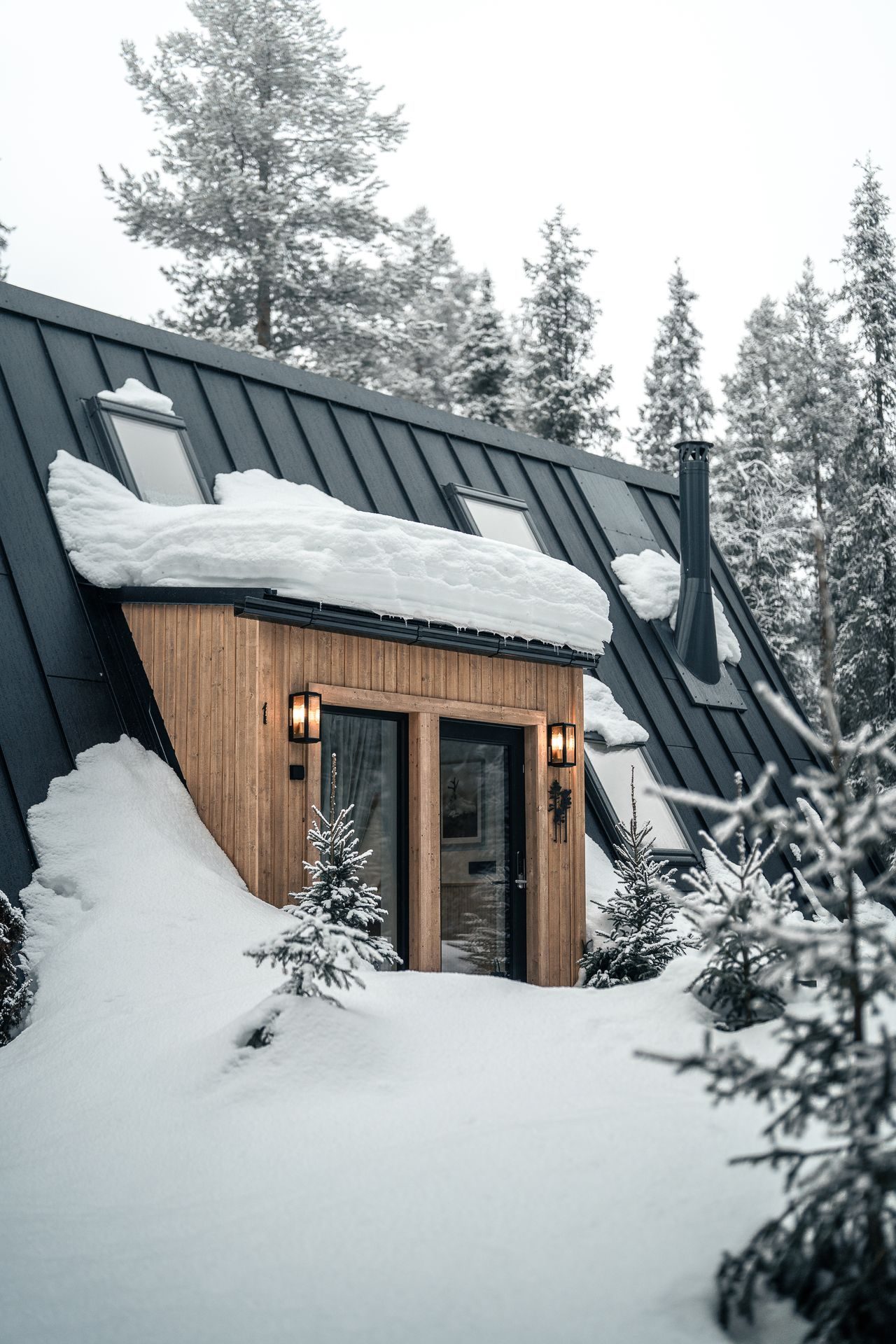The entrance of a forest cabin at Sixty Eight North beneath a snow-laden roof, lanterns lit on either side of the door and small spruce saplings dressed in snow.
