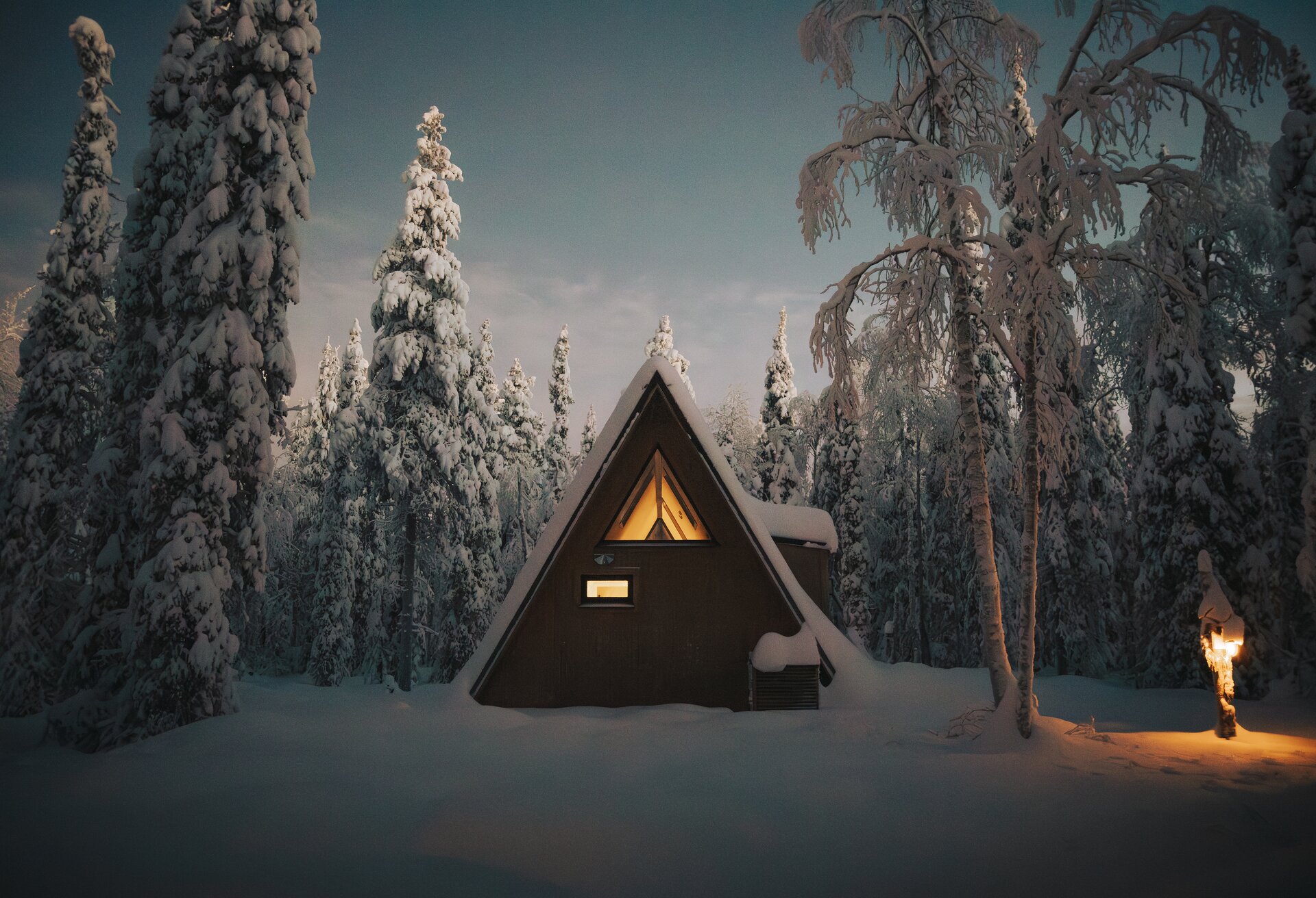 A forest cabin glowing warm at blue hour, snow-laden pines all around and a lantern lit on the path.