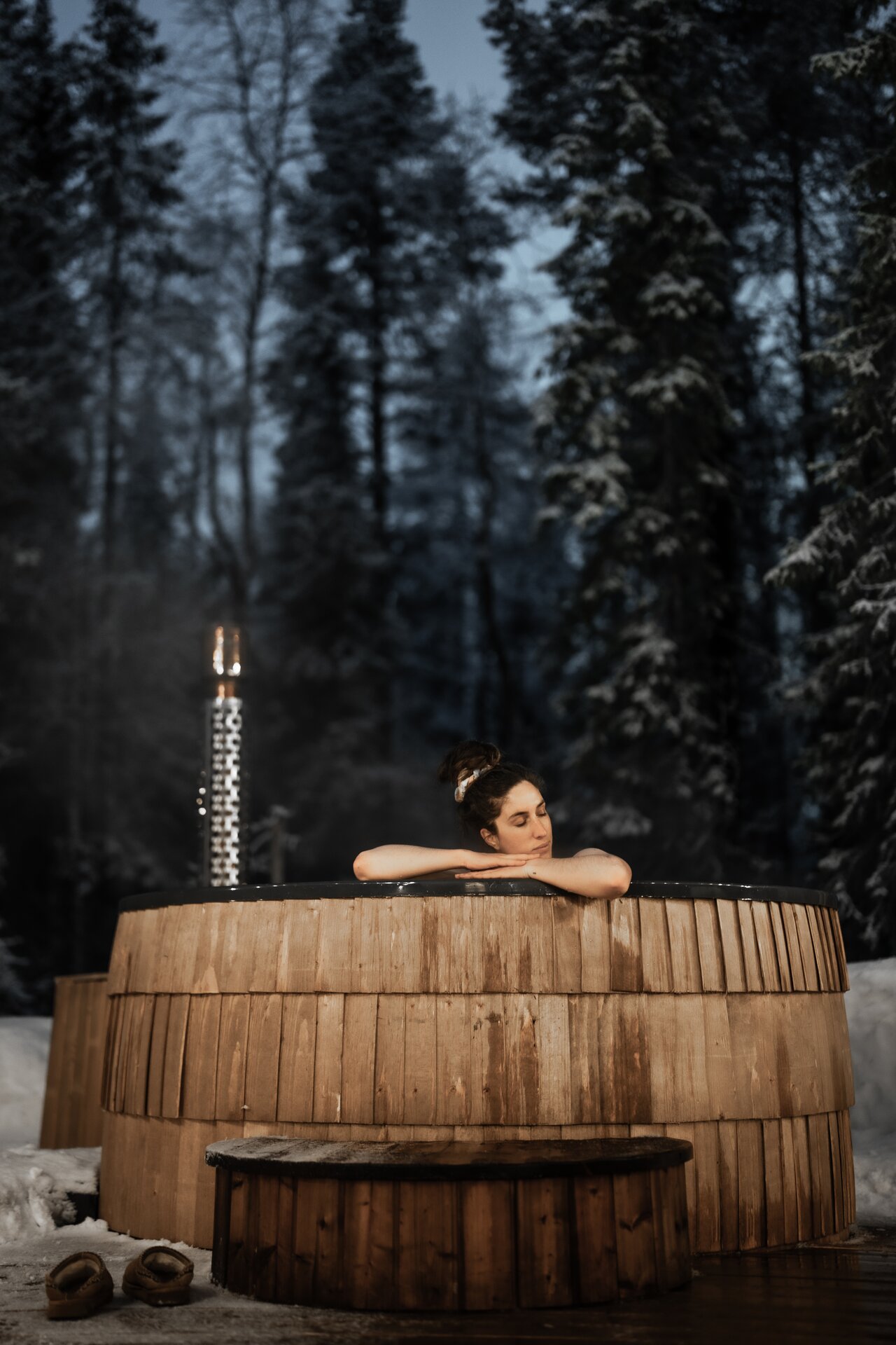 A guest resting her arms on the rim of the wooden hot tub at Sixty Eight North at dusk, tall pines and a pale sky behind.